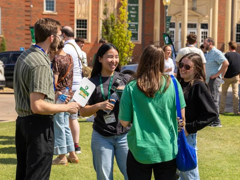 A smiling student wearing a green lanyard holds a white paper fan with the Bison logo while chatting with a small group on a sunny campus lawn. Other visitors stand in conversation behind them, with brick campus buildings in the background.