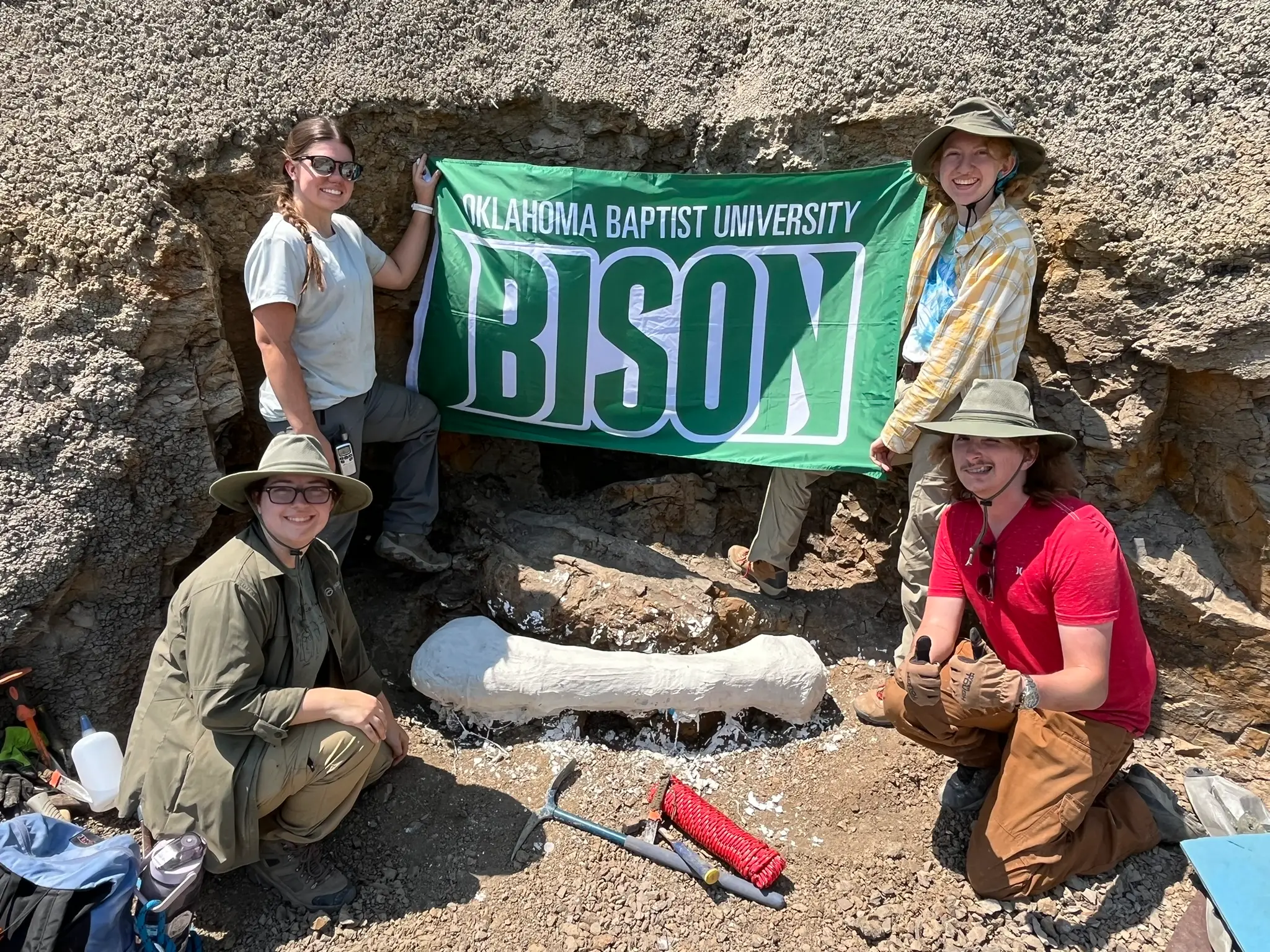 Students on a Dino Dig 2 students are in front of a large dinosaur bone. 2 more students are holding a green Oklahoma Baptist University Bison flag