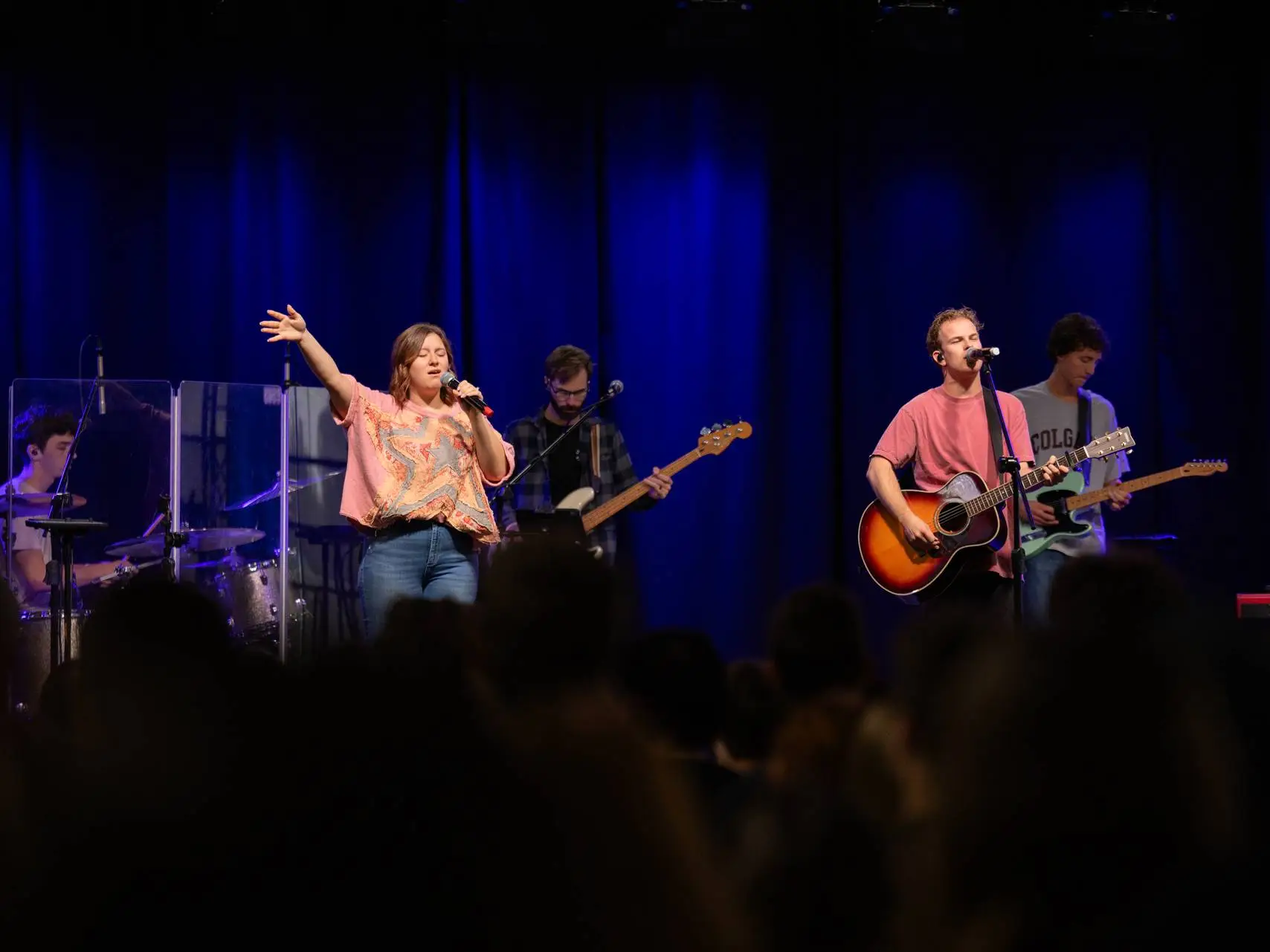 A live band performs worship music on a stage with a deep blue curtain backdrop. A female vocalist in a pink shirt has one hand raised while singing into a microphone. Beside her, a male musician plays an acoustic guitar and sings, accompanied by a drummer, bassist, and electric guitarist. The silhouette of an audience is visible in the foreground.