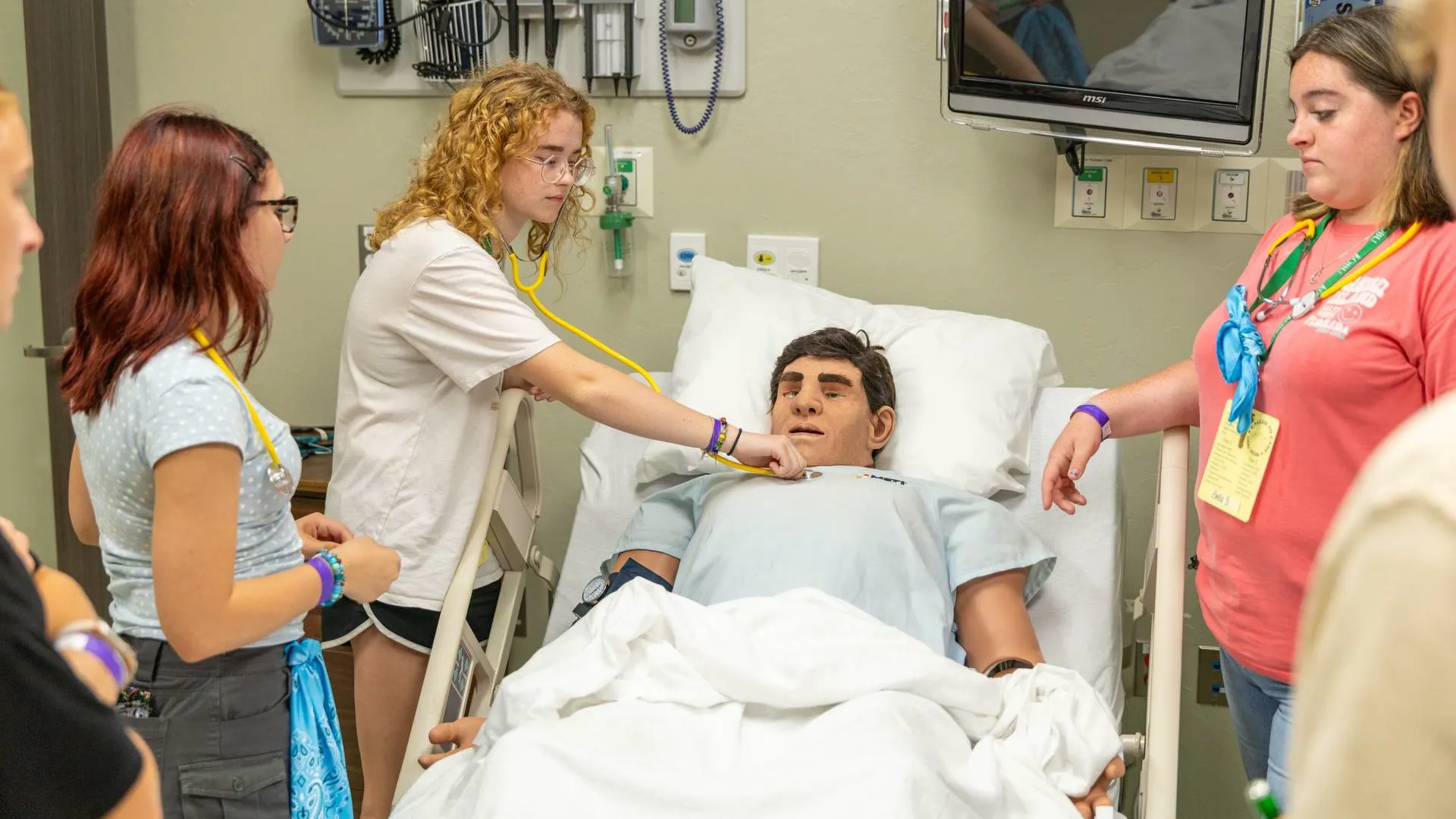 A nursing camp student with curly hair and glasses uses a stethoscope to listen to the chest of a medical training mannequin lying in a hospital bed, while two other students look on and a wall-mounted monitor and medical equipment are visible in the background exam room.