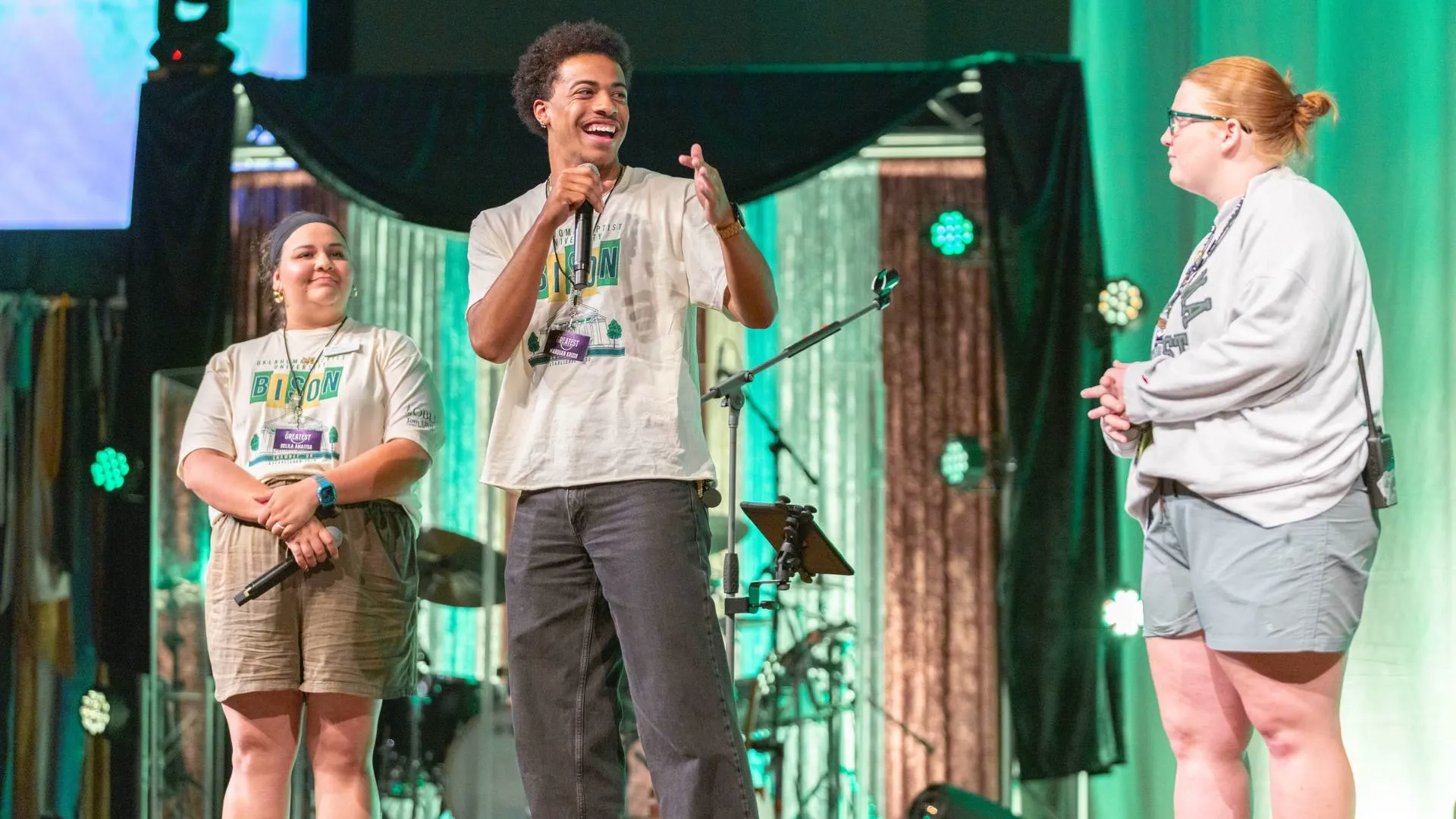 A young man on stage holds a microphone and laughs while gesturing with his free hand, wearing a cream "Oklahoma Baptist University Bison" t-shirt and a lanyard badge. A woman in a headband stands to his left and another woman with glasses stands to his right, both smiling, against a green-lit stage backdrop with draped curtains and a drum kit.