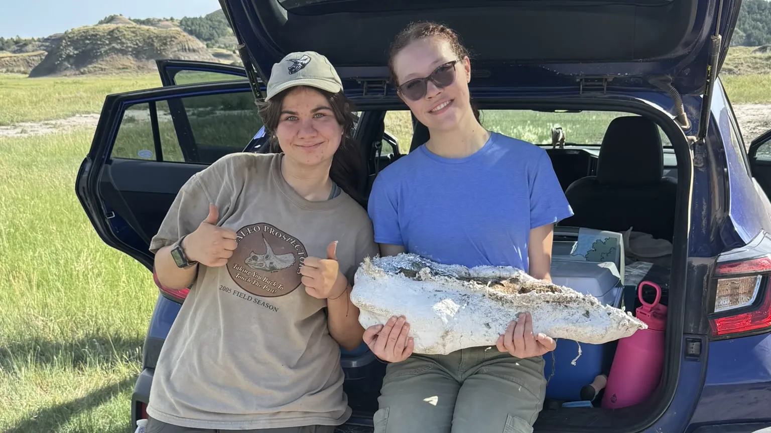 wo smiling women pose in front of an open SUV hatchback in a grassy field with badlands hills in the background. The woman on the left wears a ballcap and gives a thumbs-up, while the woman on the right, in sunglasses, helps hold up a large fossilized dinosaur jaw bone between them.