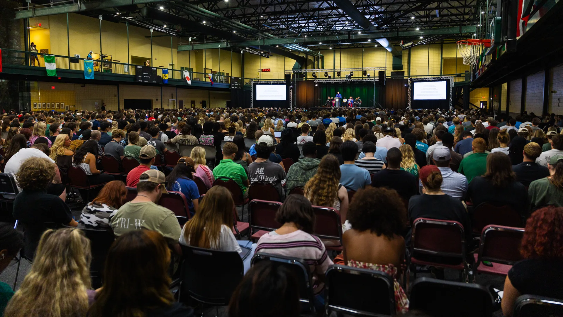 A wide view of a packed gymnasium during convocation, with rows of students seated facing a stage where speakers stand at a podium flanked by projection screens, and international flags hanging from the upper level.