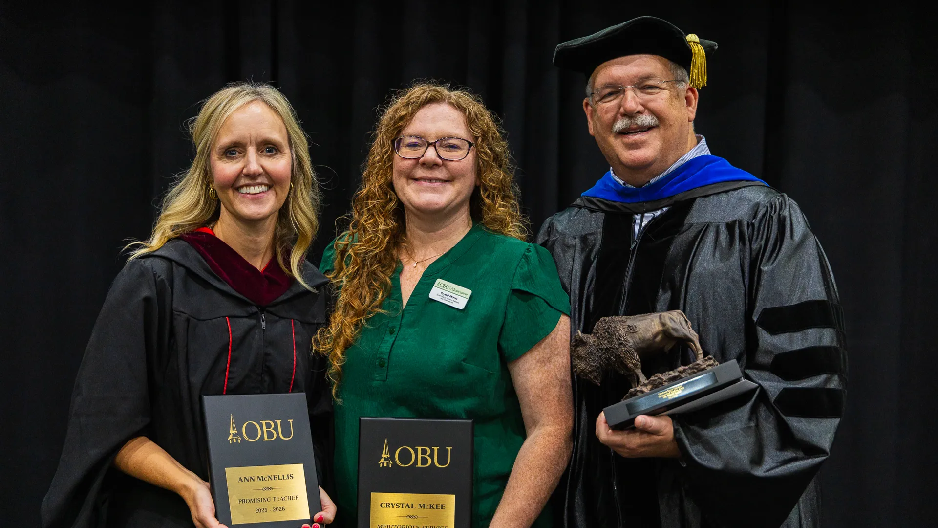 Two women and a man in academic regalia pose on stage holding OBU award plaques and a bronze bison trophy, in front of a black curtain backdrop.