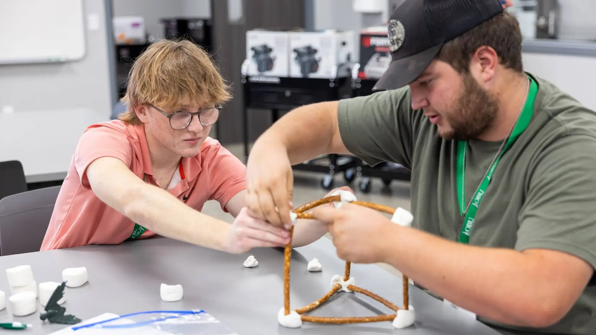 Two students at OBU's STEM Day build a geometric structure out of pretzel rods and marshmallows, working together to connect the joints of a triangular frame on a classroom table scattered with extra marshmallows.