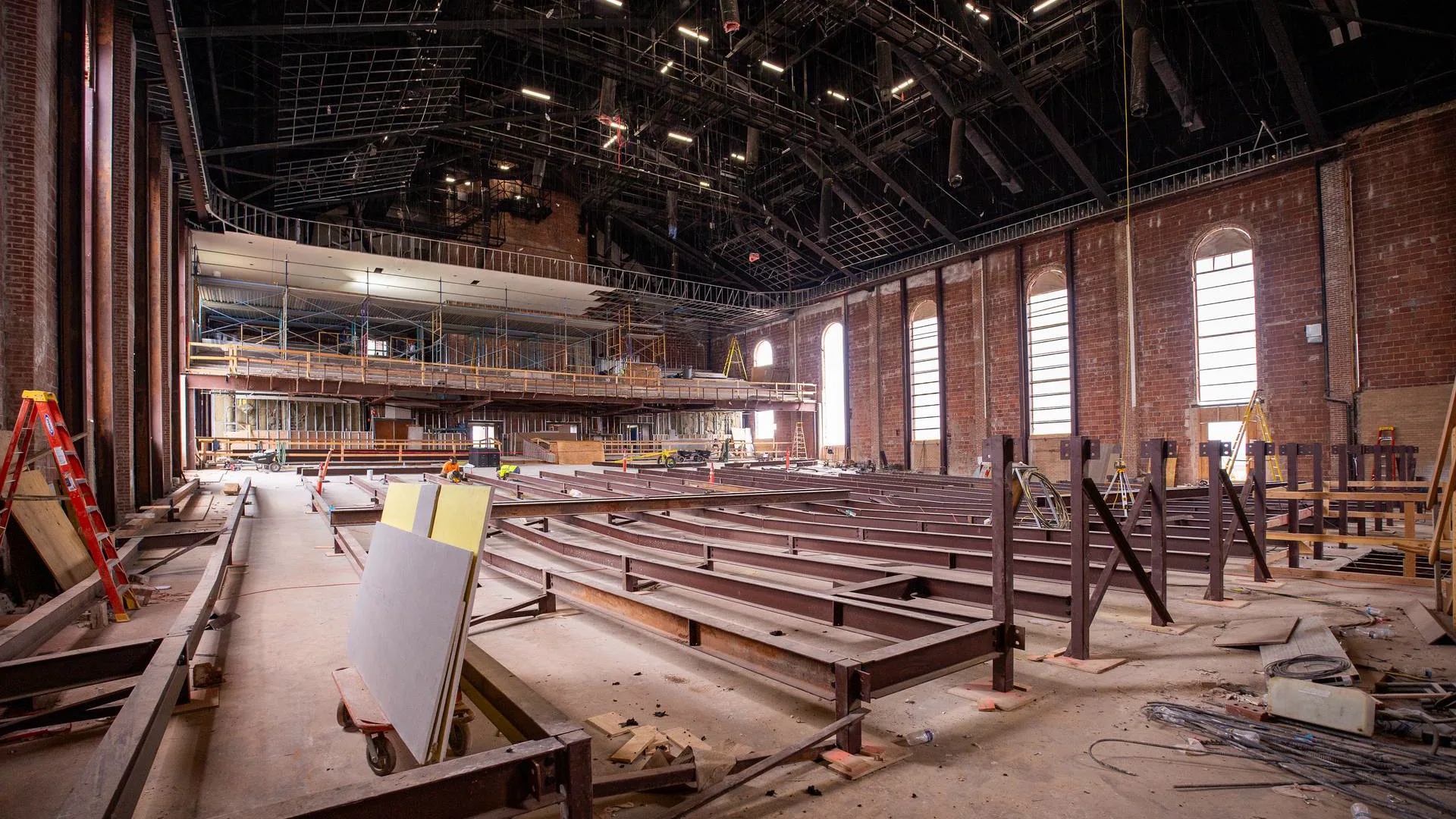 Interior of the chapel under renovation, showing rows of new steel floor framing on the main level, exposed brick walls with arched windows, and scaffolding along the upper balcony.