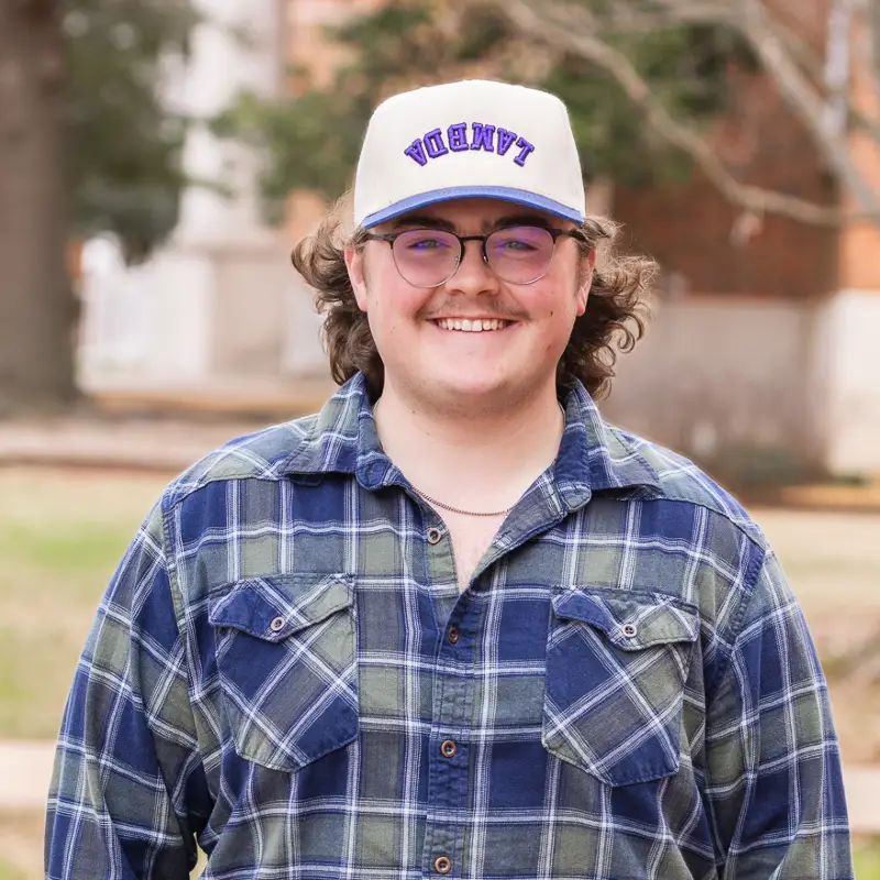 Ayden Jones. Male student in a blue and gray flannel button down tee-shirt, ball cap and glasses.