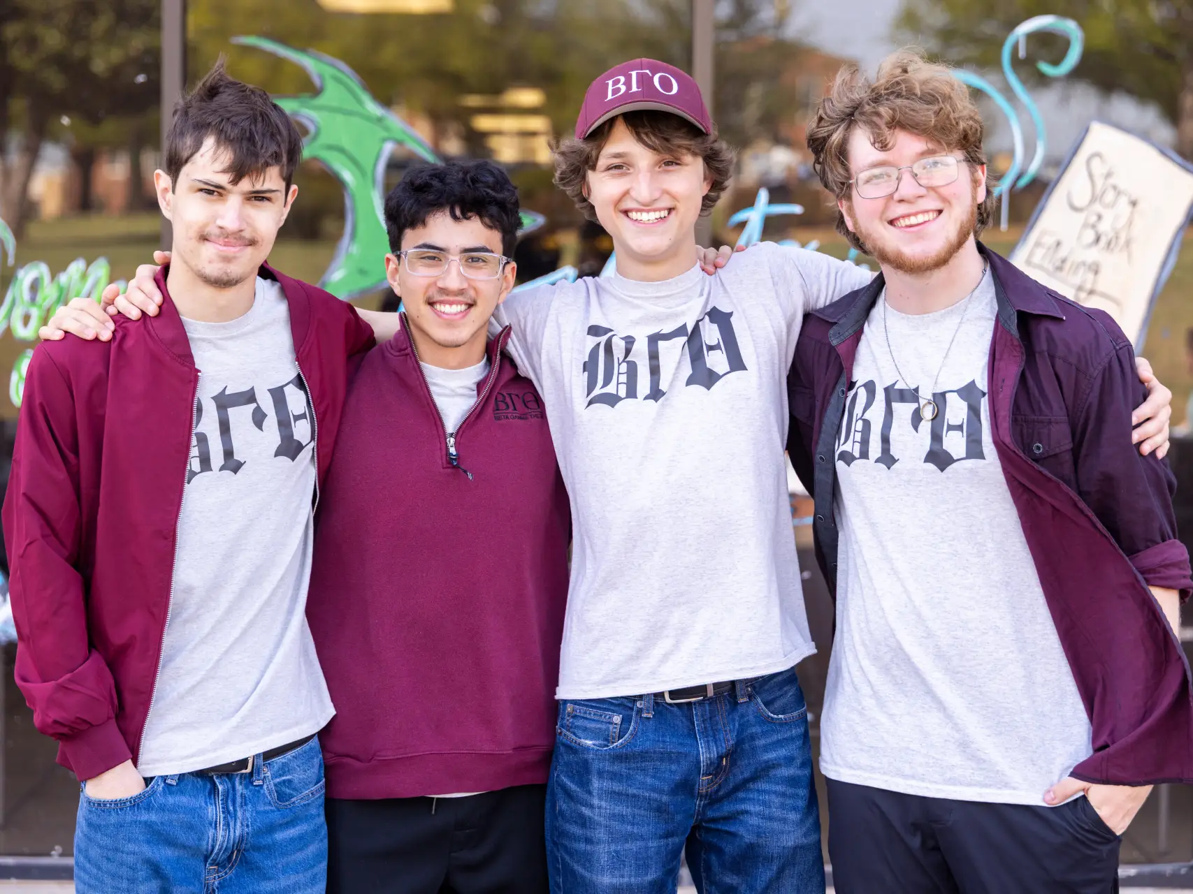 Four young men standing in front of a glass storefront, smiling for a group photo. They are wearing grey and maroon shirts featuring the Greek letters "ΒΓΘ" (Beta Gamma Theta). One man in the center is wearing a maroon baseball cap with the same letters. The background shows window art, including a green dragon and a sign that reads "Story Book Ending."