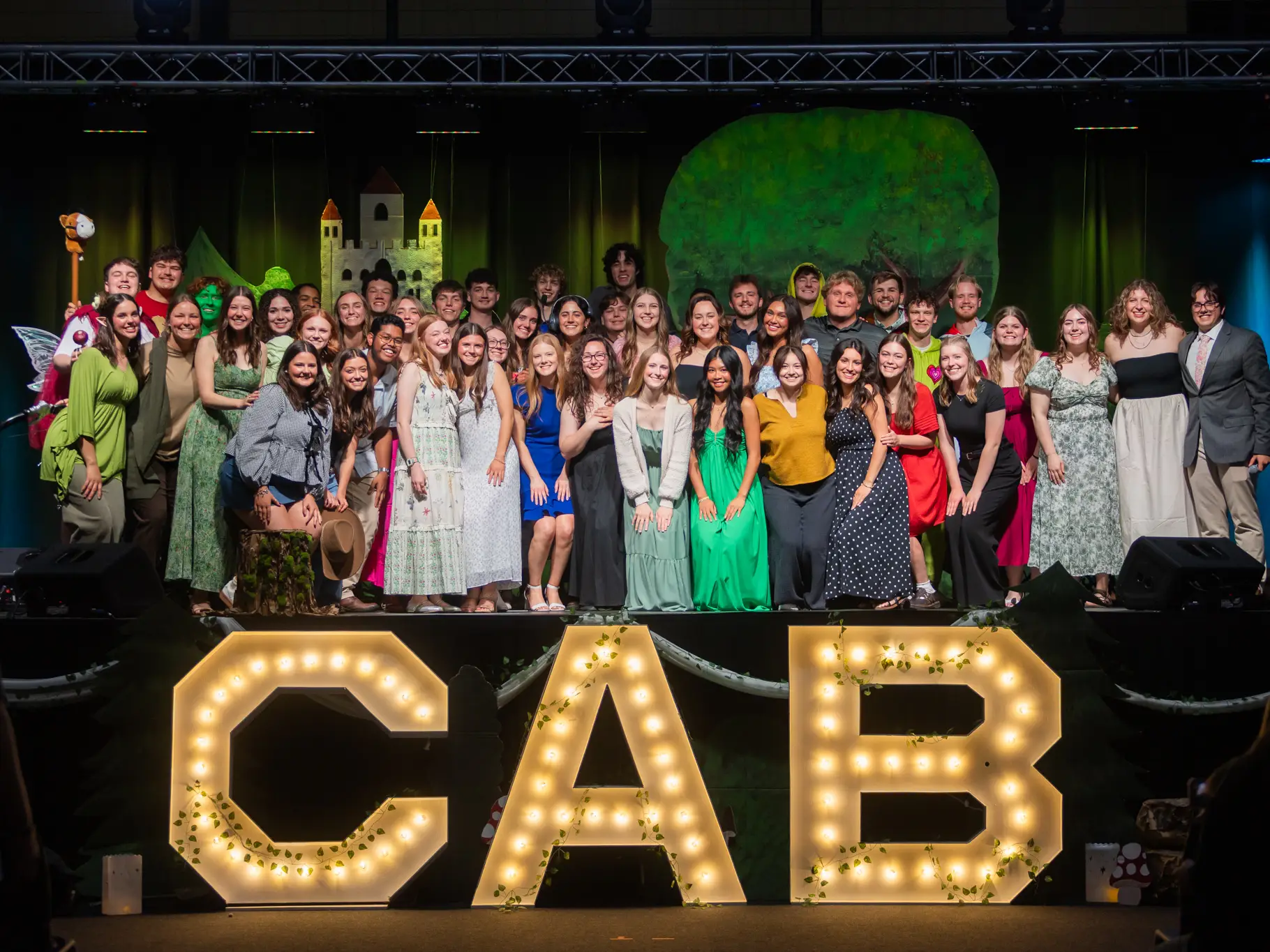 A large group of diverse college students posing on a stage. They are standing behind three large, glowing marquee letters that spell "CAB." The stage backdrop features a whimsical, hand-painted castle and a large green tree.