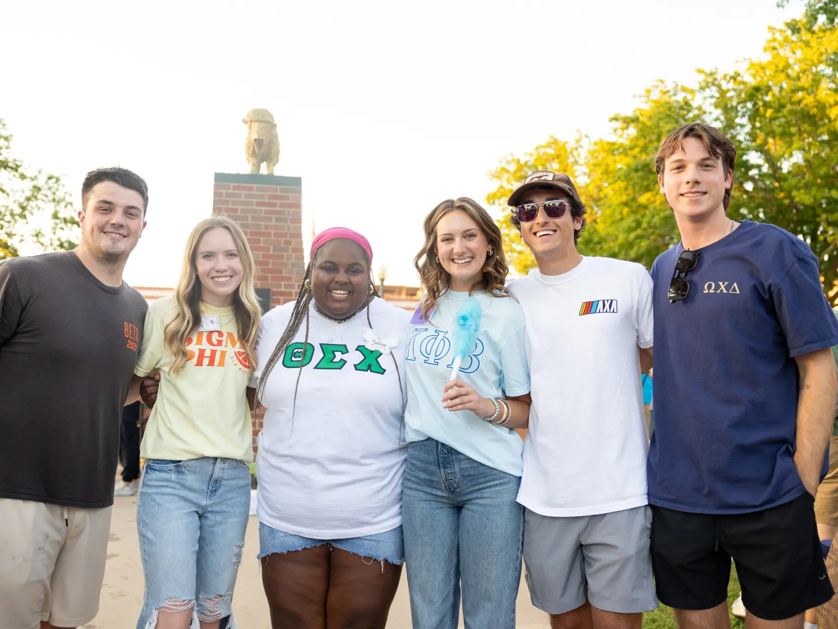 Six smiling students standing together outdoors in front of a brick pedestal. Several students are wearing t-shirts featuring Greek letters, including "Sigma Phi," "Theta Sigma Chi," and "Phi Omega Chi." One student is holding blue cotton candy.