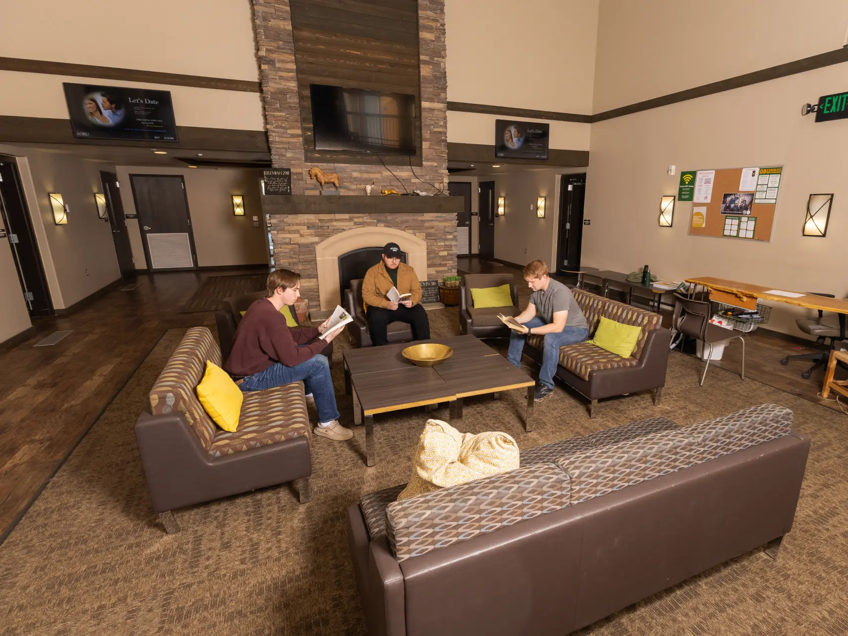 Three male students sit in a modern university common room with high ceilings and a large stone fireplace. Two students are sitting on patterned sofas and one in an armchair, all focused on reading books. The room is warmly lit with contemporary wall sconces.