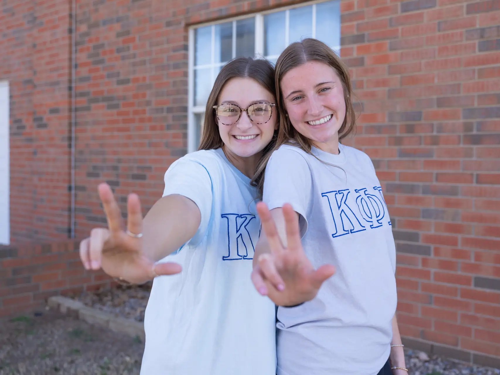 Two young women standing in front of a brick building, smiling and holding up three fingers toward the camera in a "W" shape. They are wearing t-shirts with the Greek letters "ΚΦΒ" (Kappa Phi Beta) outlined in blue. The woman on the left is wearing glasses and a light blue shirt, while the woman on the right is in a grey shirt.