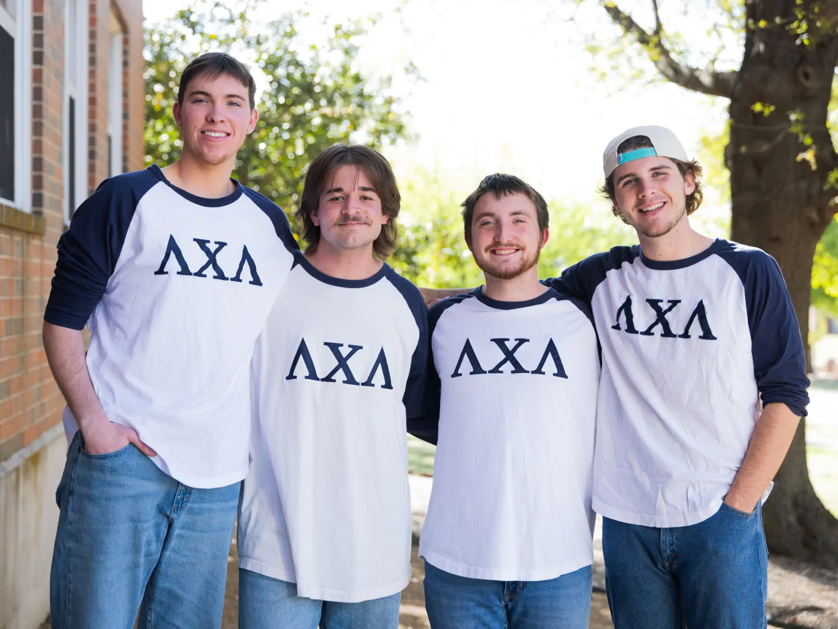 Four young men standing outside against a brick wall, smiling and posing with their arms around each other. They are wearing matching white baseball-style shirts with navy blue long sleeves. The Greek letters "ΛΧΛ" (Lambda Chi Lambda) are printed in navy blue across the chest of each shirt.