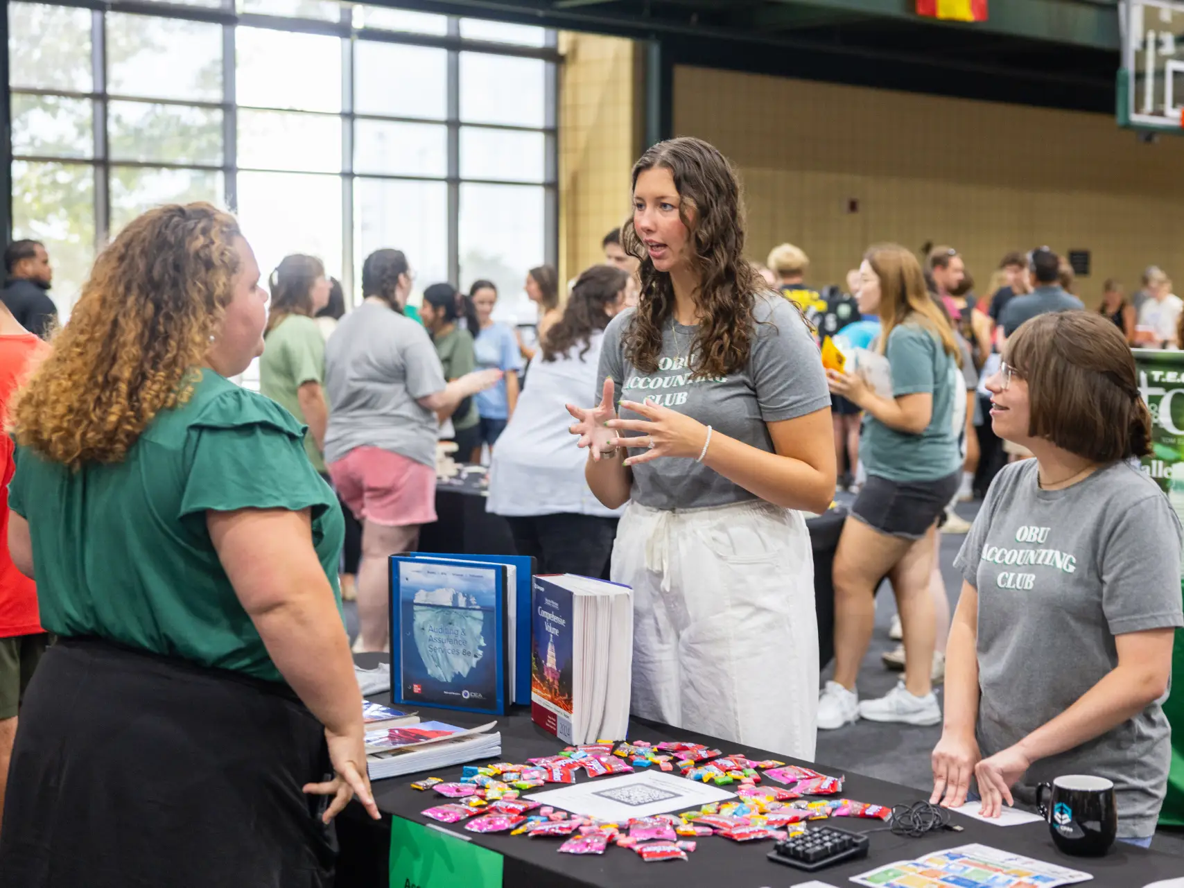 Three female students engaged in conversation at a "Student Org Fair" booth. The booth is for the "OBU Accounting Club" and is covered in candy, brochures, and a sign-up sheet. The room is filled with other students and various club tables.