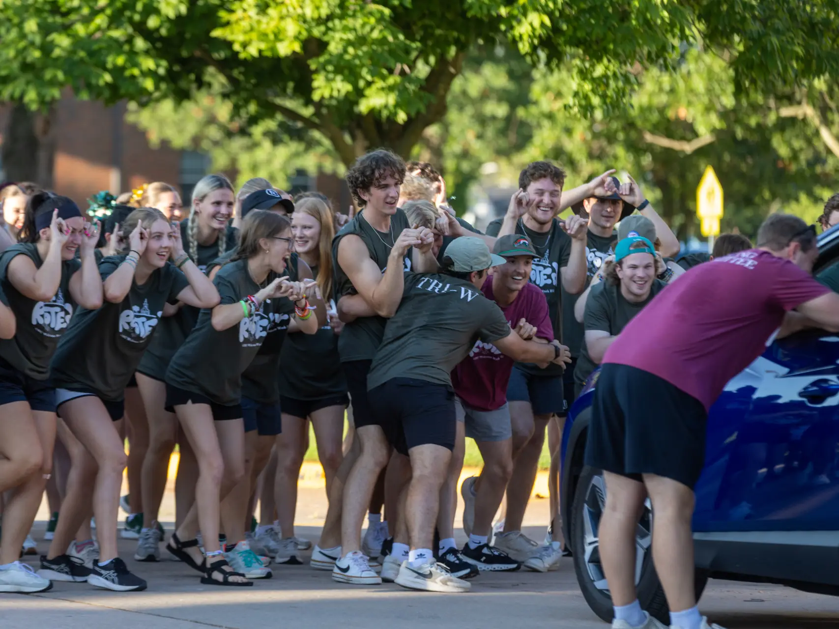 A spirited group of students wearing matching dark green "Tri-W" t-shirts. They are cheering, laughing, and making hand gestures as they surround a dark blue vehicle on a campus street.