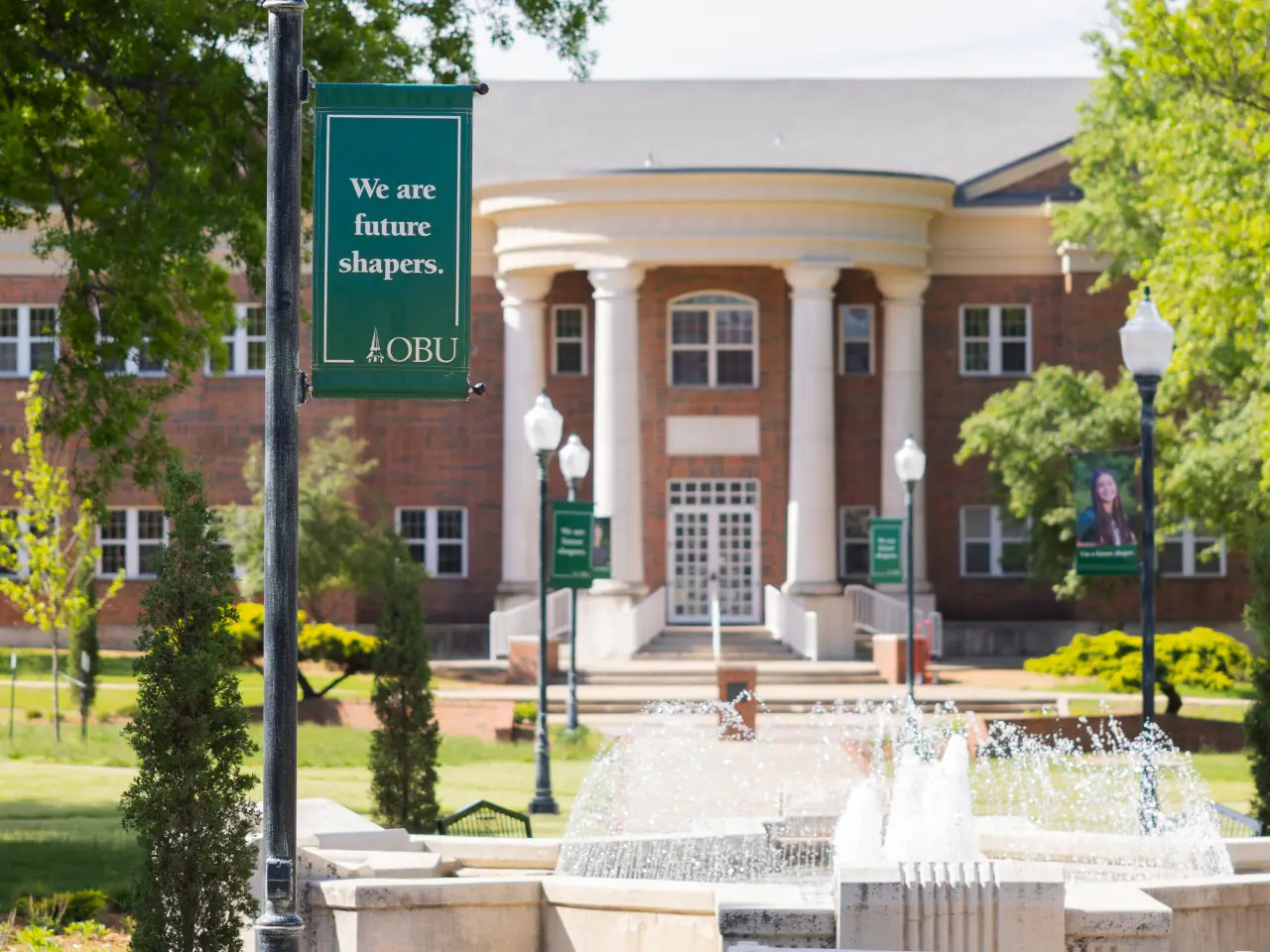A scenic view of a brick university building with white columns, framed by green trees. In the foreground, a banner on a lamp post reads, "We are future shapers. OBU." A stone fountain with spraying water is visible at the bottom of the frame.