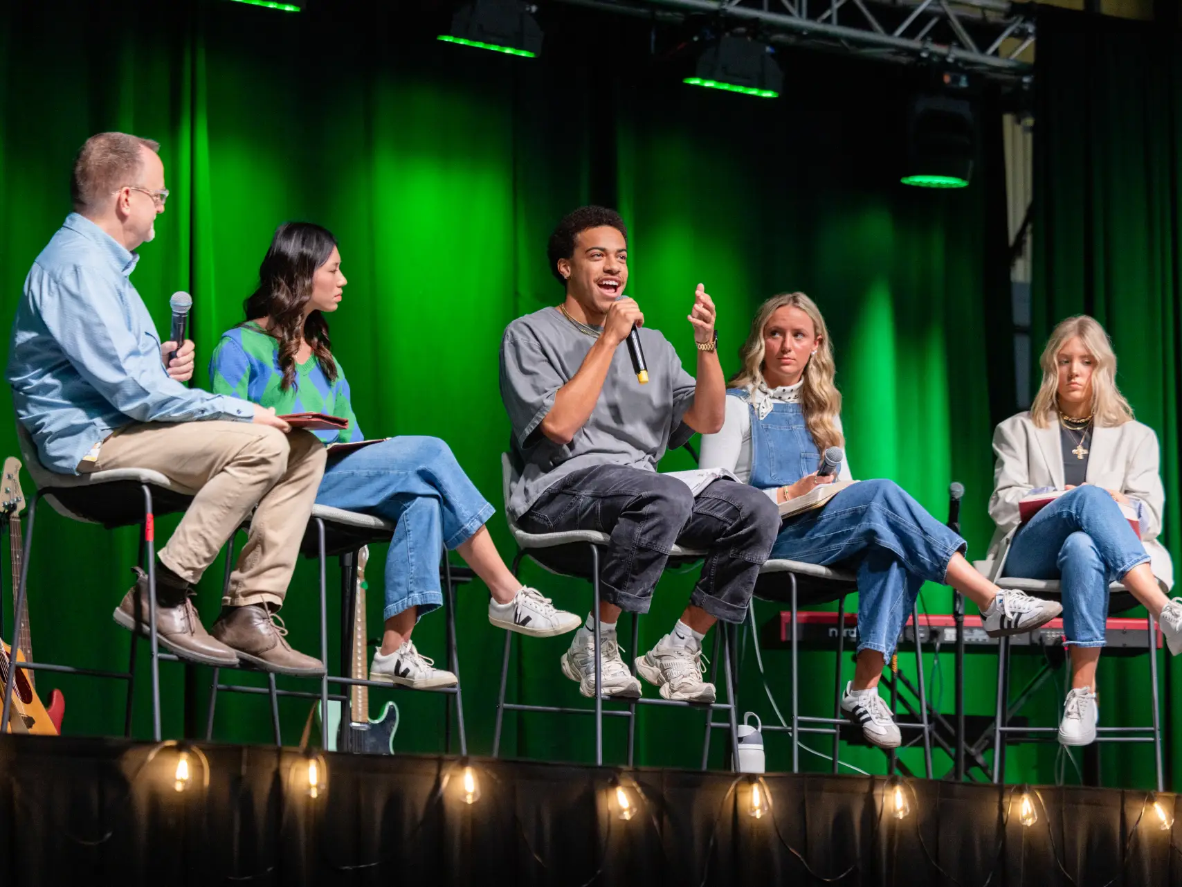 Five people sit on high stools on a stage during a panel discussion. A young man in the center is speaking into a microphone with animated hand gestures. The stage has a vibrant green curtain backdrop and is lit with warm string lights along the front edge.
