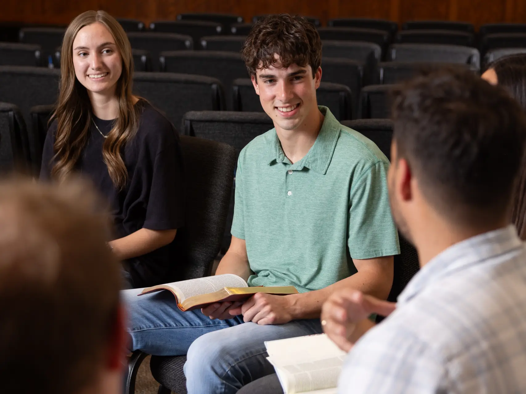 A young man in a green polo shirt sits in a dark auditorium, smiling and holding an open book during a group discussion. A young woman to his left listens intently, while the back of another person’s head is visible in the foreground.