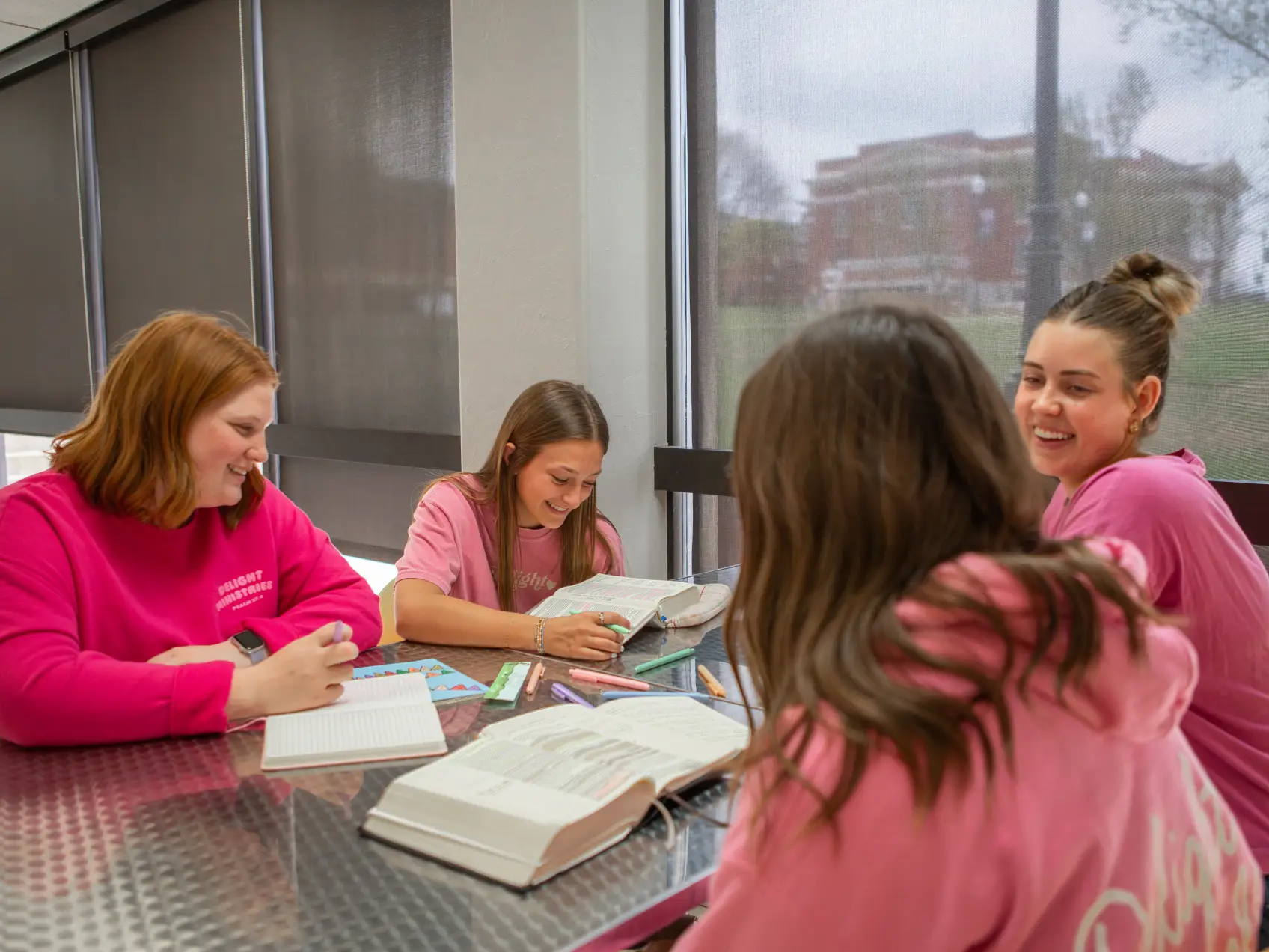 Four young women, all wearing different shades of pink shirts and sweatshirts, sit around a metal table in a bright room with large windows. They are smiling and laughing while studying together, with open Bibles and notebooks spread out on the table.