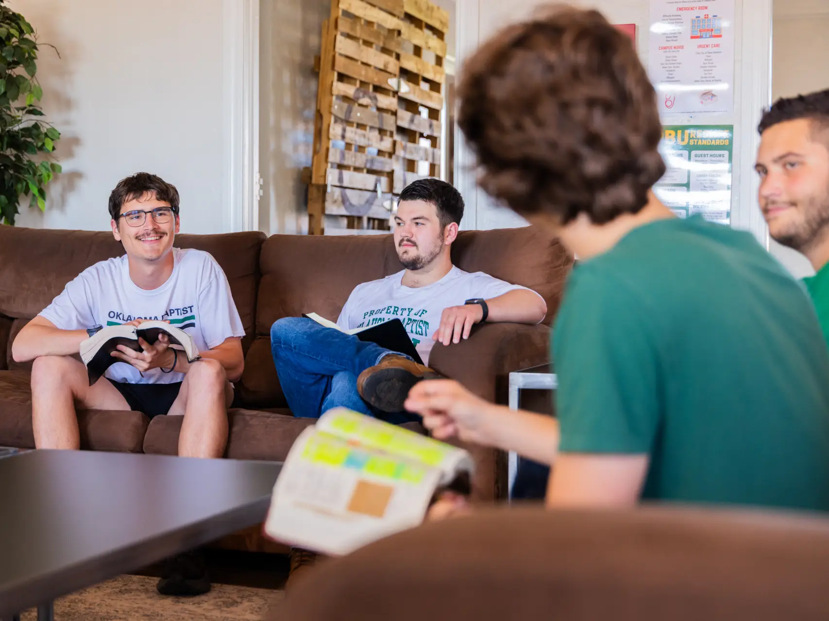 Four young men sit in a casual dorm common area. Two are sitting on a brown sofa holding open Bibles, while another student in a green shirt is blurred in the foreground, gesturing toward his own Bible during a discussion.
