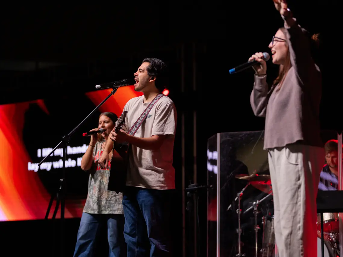 An image shows three people singing on a stage against a dark background. In the foreground on the right, a woman in a grey sweater and tan pants sings into a microphone with one arm raised. In the center, a man in a light-colored t-shirt and jeans plays an acoustic guitar and sings into a microphone on a stand. To his left and slightly behind him, another woman in a patterned shirt and jeans also sings into a microphone. Behind them, a large screen displays red abstract graphics and white text. To the far right, a drummer is partially visible behind a clear plastic screen.