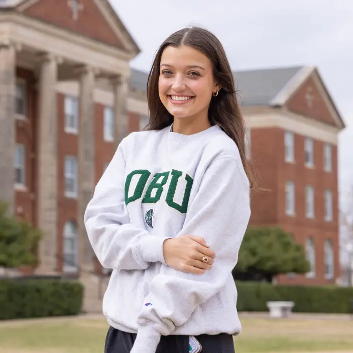 A portrait of a smiling young woman, Katie Mayes, standing outdoors on a college campus. She is wearing a white "OBU" sweatshirt and has her arms folded. Large brick academic buildings are visible in the background.