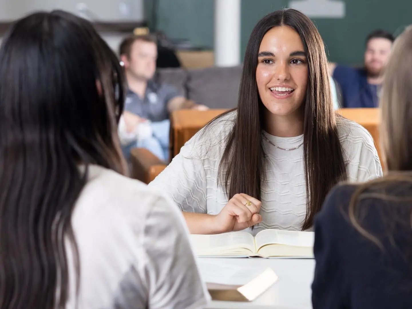 A young woman with long dark hair smiles and speaks during a small group study. She is seated at a white table with an open book in front of her, engaged in conversation with others who are partially visible.