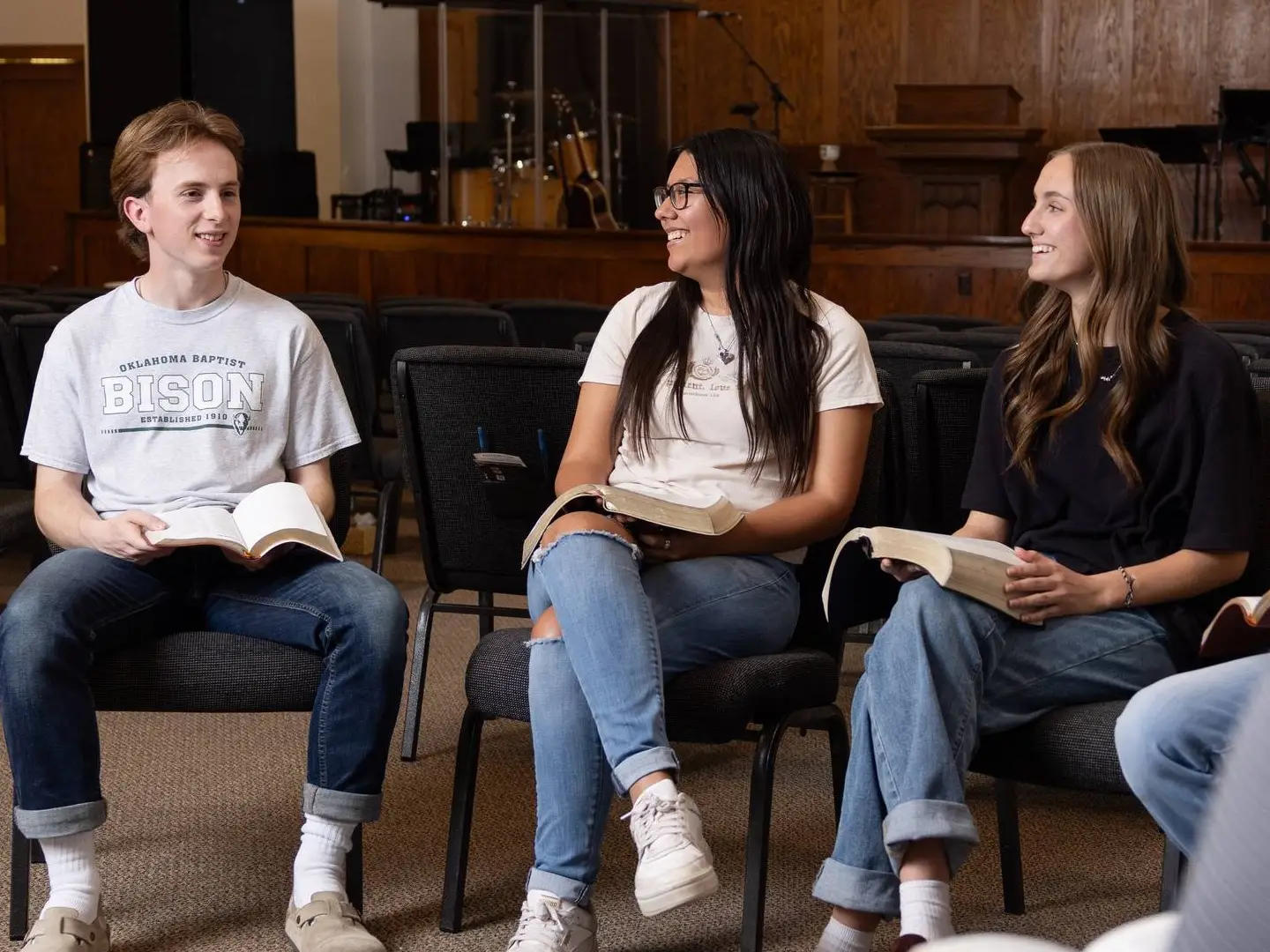 Three students sit in a circle of chairs in a room with wood-paneled walls. They each have an open Bible on their lap and are smiling and laughing together during a group discussion. One student wears a grey "Bison" T-shirt.