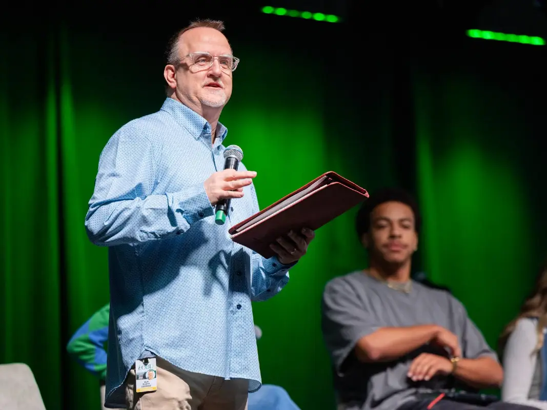 Man with glasses and a light blue patterned button-down shirt stands on a stage, holding a microphone and a brown leather folder. He appears to be speaking or leading an event against a vibrant green curtain backdrop. In the background, a younger man in a grey t-shirt sits and listens, partially out of focus.