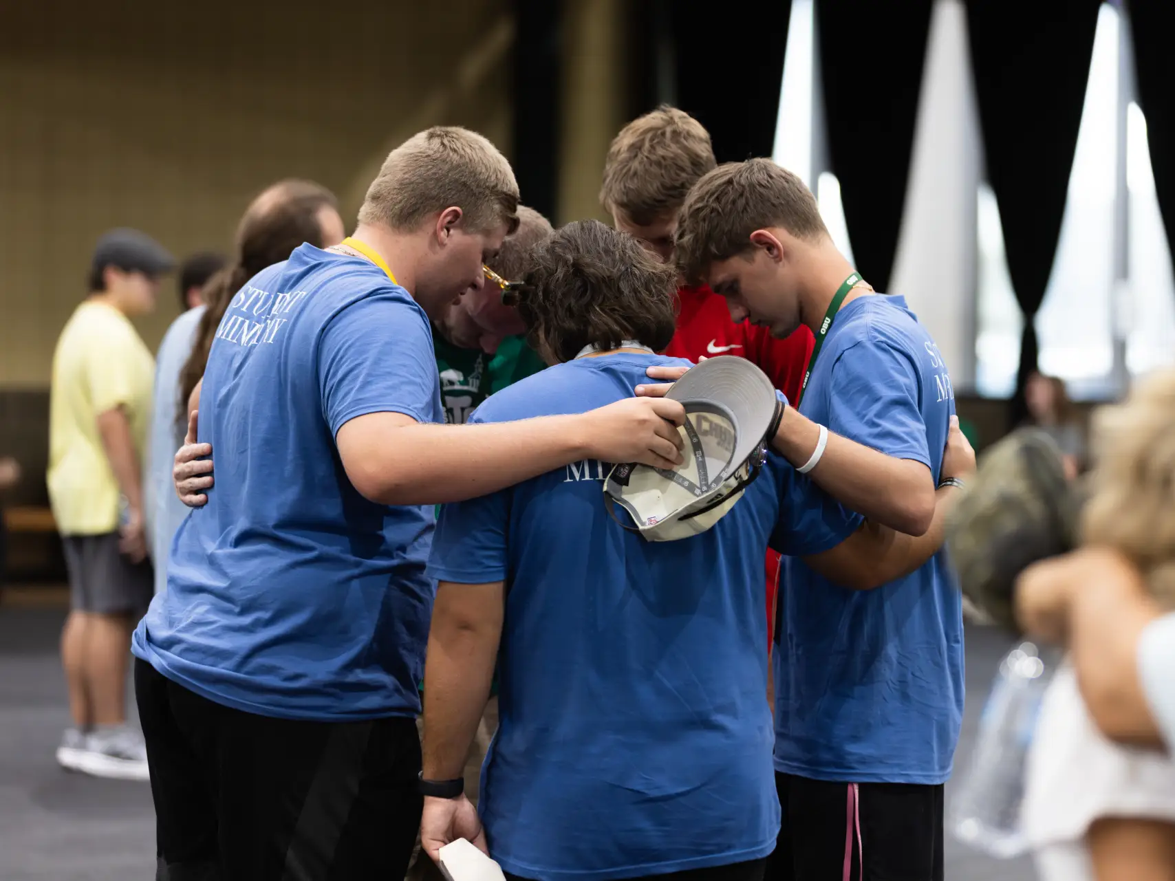 A group of five young men stand in a close circle, their heads bowed and arms around each other's shoulders in a moment of quiet solidarity or prayer. Three of the young men wear bright blue t-shirts; one shirt has "STUDENT MINISTRY" printed on the back. A fourth young man, partially visible, wears a red shirt. In the background, other people and large white fabric panels are softly blurred, keeping the focus on the intimate huddle of the group.